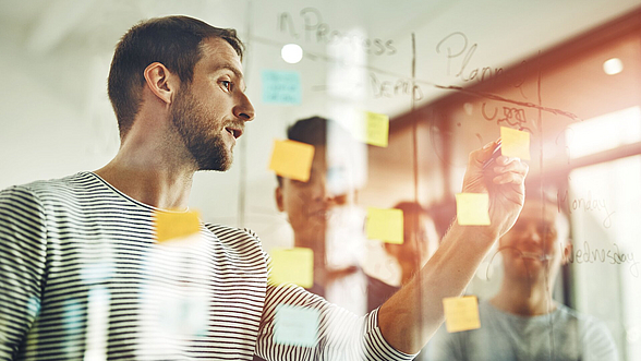 © PeopleImages -istock.com Ein Mann steht an einer Wand und beschriftet Klebezettel.