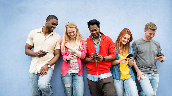 © franckreporter – istock.com Junge Menschen stehen an einer Wand und haben Handys in der Hand
