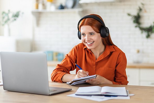 Eine junge Frau sitzt lächelnd mit einem Headset und einem Notizblock vor einem Laptop.