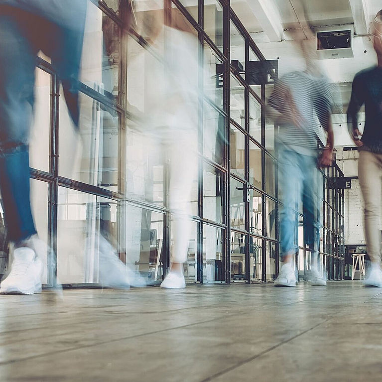 Unterschiedliche Menschen gehen durch einen Büroflur und man sieht sie in Bewegung.